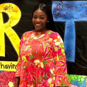 A woman wearing a vibrant floral dress stands in front of a colorful, decorated backdrop. She is smiling and looking at the camera.