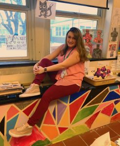 A woman in a pink top and red pants sits on a colorful ledge in a classroom, surrounded by various art pieces and papers. She smiles at the camera with one leg resting on the ledge.