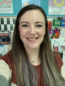 A woman with long brown hair and a reddish-brown sweater smiles at the camera. Behind her, colorful decorations and motivational posters are visible on the wall.